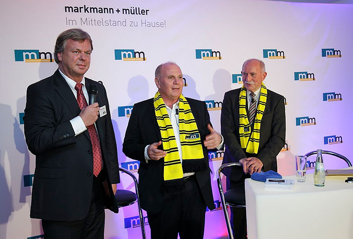 Uli Hoeneß, langjähriger Vorstand und Präsident des FC Bayern, Sportreporter Werner Hansch und m+m Geschäftsführer Michael Müller in einem Podiumsgespräch vor einer m+m Logowand.